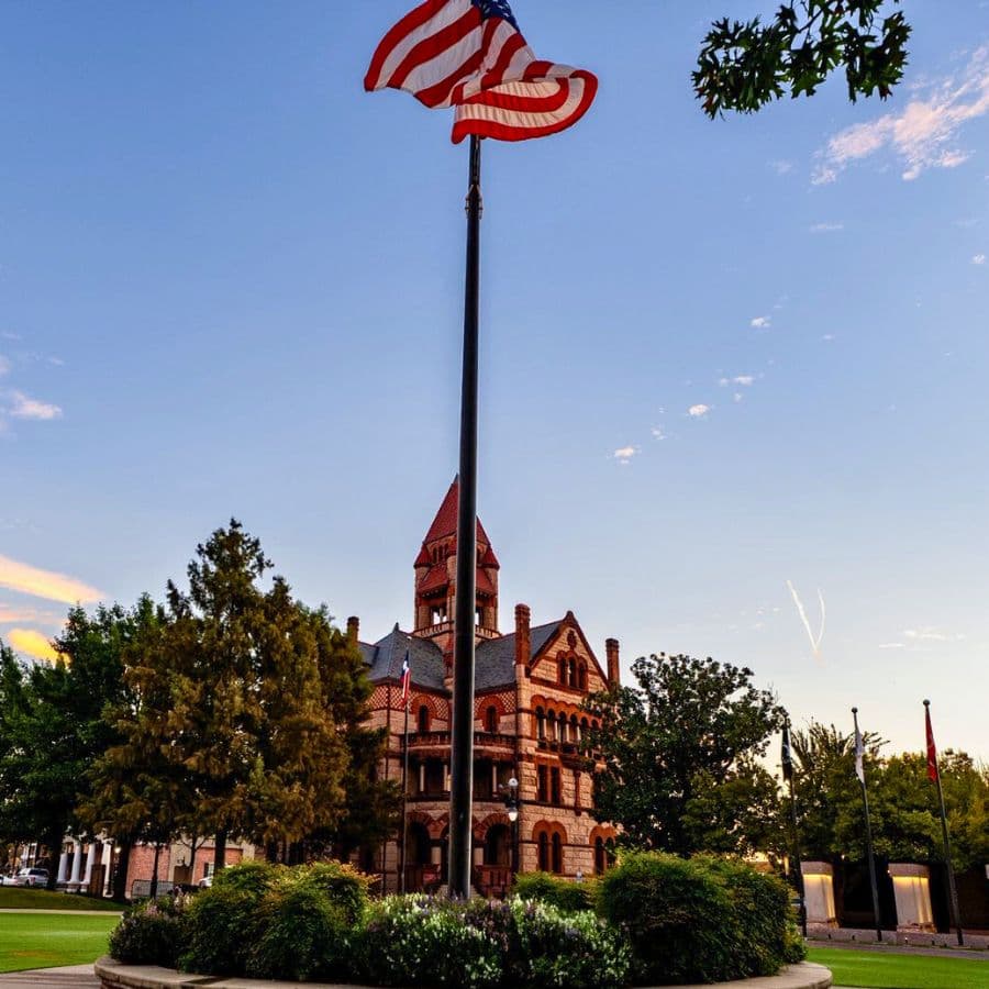 A historic red brick building with a tall flagpole displaying the American flag amidst green trees and a blue sky.