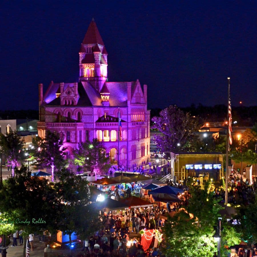 A historic building illuminated in purple stands over a bustling evening market filled with people and tents.